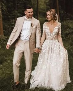 a man and woman holding hands walking through the woods in their wedding dresses with flowers on them