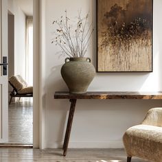 a vase sitting on top of a wooden table in front of a painting and chair