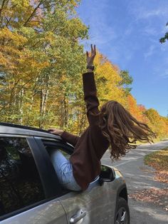 a woman is waving out the window of a car on a road with trees in the background