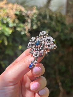 A literal bouquet of jewels! This stunning Sapphire and Diamond brooch is a feast for the eyes, composed in a design reminiscent of Victorian era Giardinetti (little gardens) jewelry. Crafted in antique 14 carat gold, this brooch features a dazzling array of 78 miniature bezel-set diamonds, arranged in a botanical motif. Dotted along the corners of the brooch are 7 round mix cut emeralds, which add a whimsical flair to the silhouette. The centerpieces of this adornment however are the two vibrant blue cabochon-cut sapphires: the first is nestled in the very center of the brooch, surrounded by a halo of diamonds, while the second dangles at the bottom like an elegant flower bulb. A perfect mix of glamour and lushness, this treasure is the perfect addition to any antique jewelry collection! Antique Baroque Brooches With 17 Jewels, Ornate Baroque Jewelry With Rose Cut Diamonds, Elegant Diamond Multi-stone Brooch, Ornate Rose Cut Diamond Brooches For Weddings, Antique Diamond Brooch Jewelry, Ornate Diamond Brooch For Anniversary, Ornate Diamond Brooch As Gift, Ornate Diamond Brooch Gift, Ornate Diamond Brooches For Anniversary