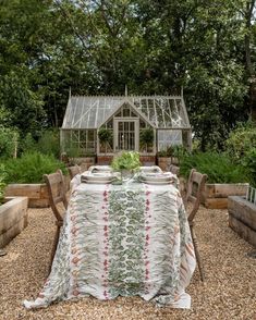 a table set for two in the middle of a garden with an old greenhouse behind it