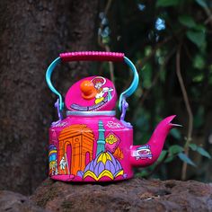 a pink teapot with colorful designs on it sitting on a rock next to a tree