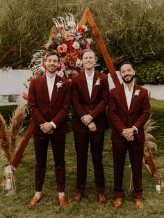 three men in maroon suits standing next to each other on the grass with an orange and red floral arrangement behind them