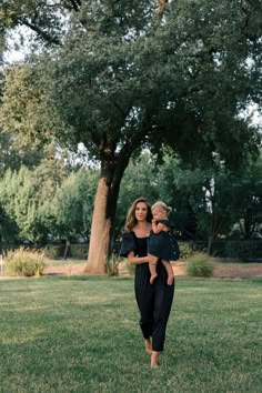 a woman holding a baby in her arms while standing on top of a lush green field