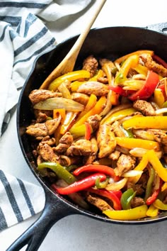 a skillet filled with chicken and peppers on top of a white table cloth next to a wooden spoon