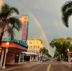 a rainbow shines in the sky over a street with buildings and palm trees on both sides