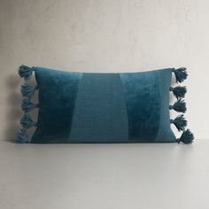 a teal pillow with tassels on the bottom and side, against a white wall