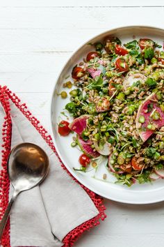 a white plate topped with salad next to a silver spoon and red napkin on top of a wooden table