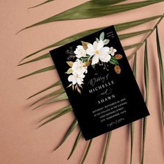 a black wedding card with white flowers and pineconi on the front, next to palm leaves