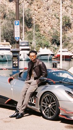 a man sitting on top of a silver sports car in front of some water and boats