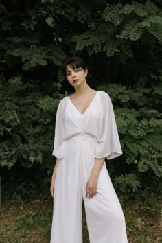 a woman standing in front of some trees wearing a white jumpsuit and black heels