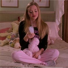 a woman sitting on a bed holding a pink teddy bear and looking at her cell phone