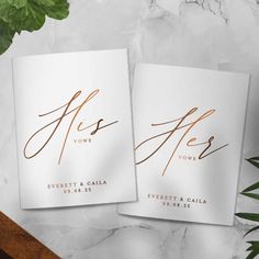 two white cards with gold foil on them sitting next to a plant and potted plant