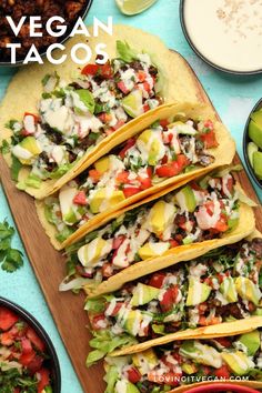 several tacos are lined up on a cutting board next to bowls of salsa and avocado