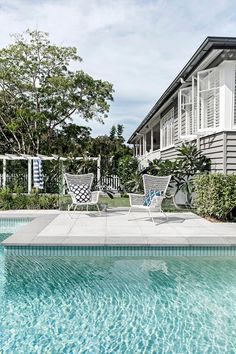 two lawn chairs sitting on the edge of a swimming pool next to a house with white shutters