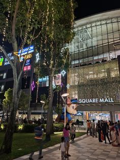 people are walking around in front of a building with lights on the sides and trees