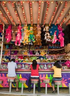 two people sitting at a carnival booth with stuffed animals on the wall and lights hanging from the ceiling