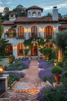 a house with lots of plants and lights in the front yard, surrounded by lavender bushes