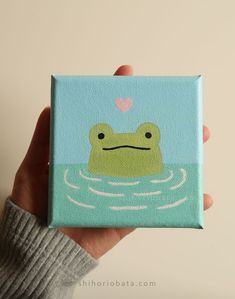 a hand holding up a small square painting with a frog on it's face