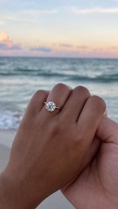 a person's hand holding an engagement ring in front of the ocean at sunset