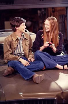 two young people sitting on the hood of a car talking to each other and laughing