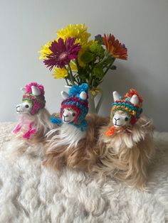 three llamas wearing knitted hats and scarves next to a vase with flowers