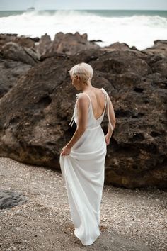 a woman standing on top of a rocky beach next to the ocean wearing a white dress