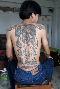 the back of a man with tattoos on his body sitting in front of a bed