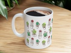 a coffee mug with cactus designs on it sitting on a wooden table next to a potted plant