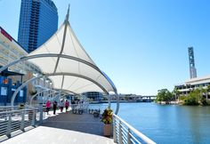 people are walking on the boardwalk next to the water and buildings in the background with white awnings over them