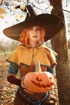 a woman dressed as a witch with a pumpkin in her hand, standing next to a tree