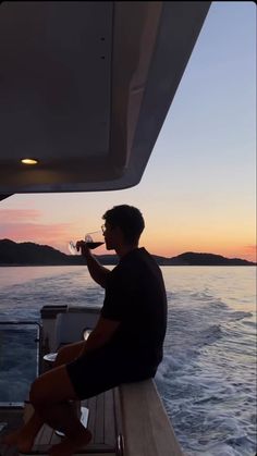 a man is sitting on the edge of a boat drinking from a wine glass at sunset