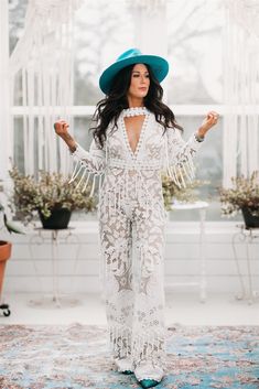 a woman standing in front of a window wearing a blue hat and white lace jumpsuit