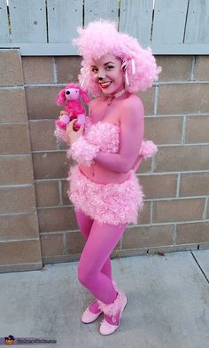 a woman dressed in pink holding a stuffed animal and smiling at the camera while standing next to a brick wall