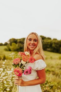 a woman in a white dress is holding some pink and orange flowers while smiling at the camera