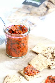 a glass jar filled with red sauce next to crackers on a wooden tablecloth