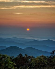 the sun setting over mountains with trees in foreground and clouds in the sky above