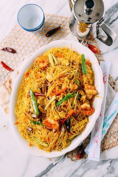 a white bowl filled with noodles and shrimp on top of a table next to a silver teapot