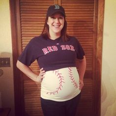 a pregnant woman is standing in front of a wooden door and wearing a baseball shirt