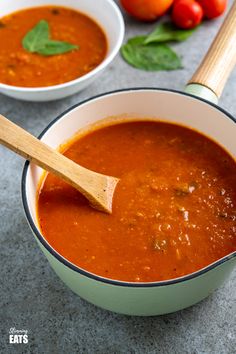 two bowls of tomato soup with basil and tomatoes in the background