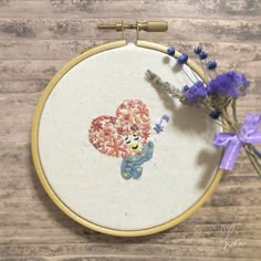 a teddy bear with flowers in it's hand on a wooden table next to a cross stitch hoop