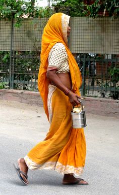 a woman walking down the street with a bucket in her hand and wearing an orange sari