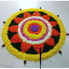 a circular flower arrangement on the ground with two black sticks in front of it and an orange, pink, yellow, and white center