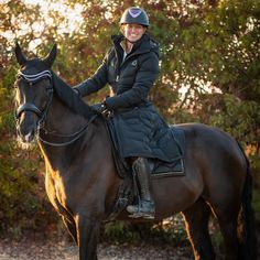 a woman riding on the back of a brown horse in front of bushes and trees