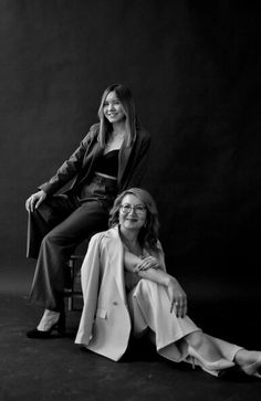 two women sitting next to each other posing for a black and white photo in front of a dark background