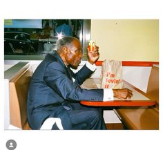 an old man in a tuxedo eating a hot dog at a restaurant table