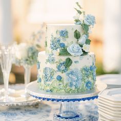 a wedding cake with blue flowers and greenery on the top is sitting on a table