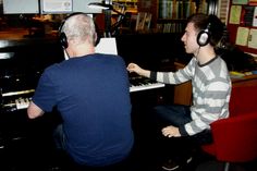 a man sitting at a piano with headphones on and an older man standing next to him