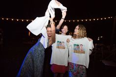 three people holding up t - shirts in the air with lights strung above them at night