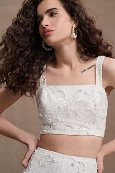 a woman in a white crop top and skirt with her hands on her hips, looking off to the side